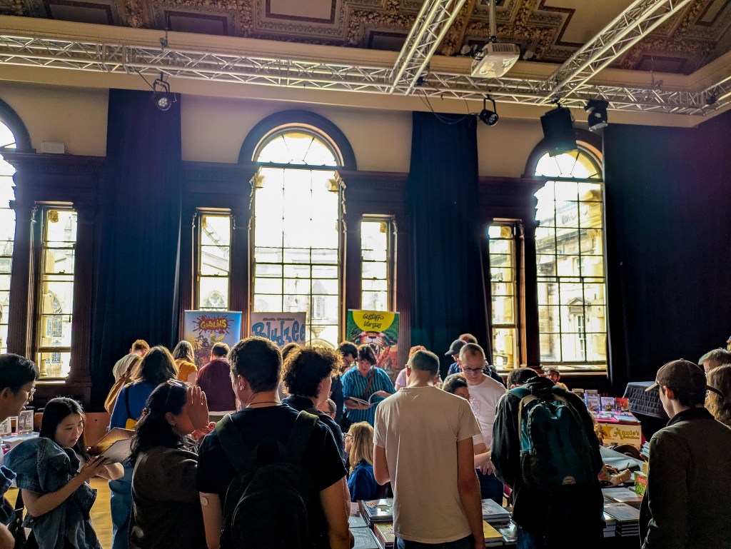 An elegant classical room with large windows. There is a crowd of people and many stalls and pull up banners