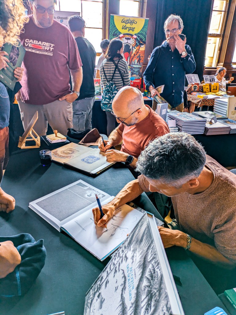 Two men sit at a table signing and sketching books.
